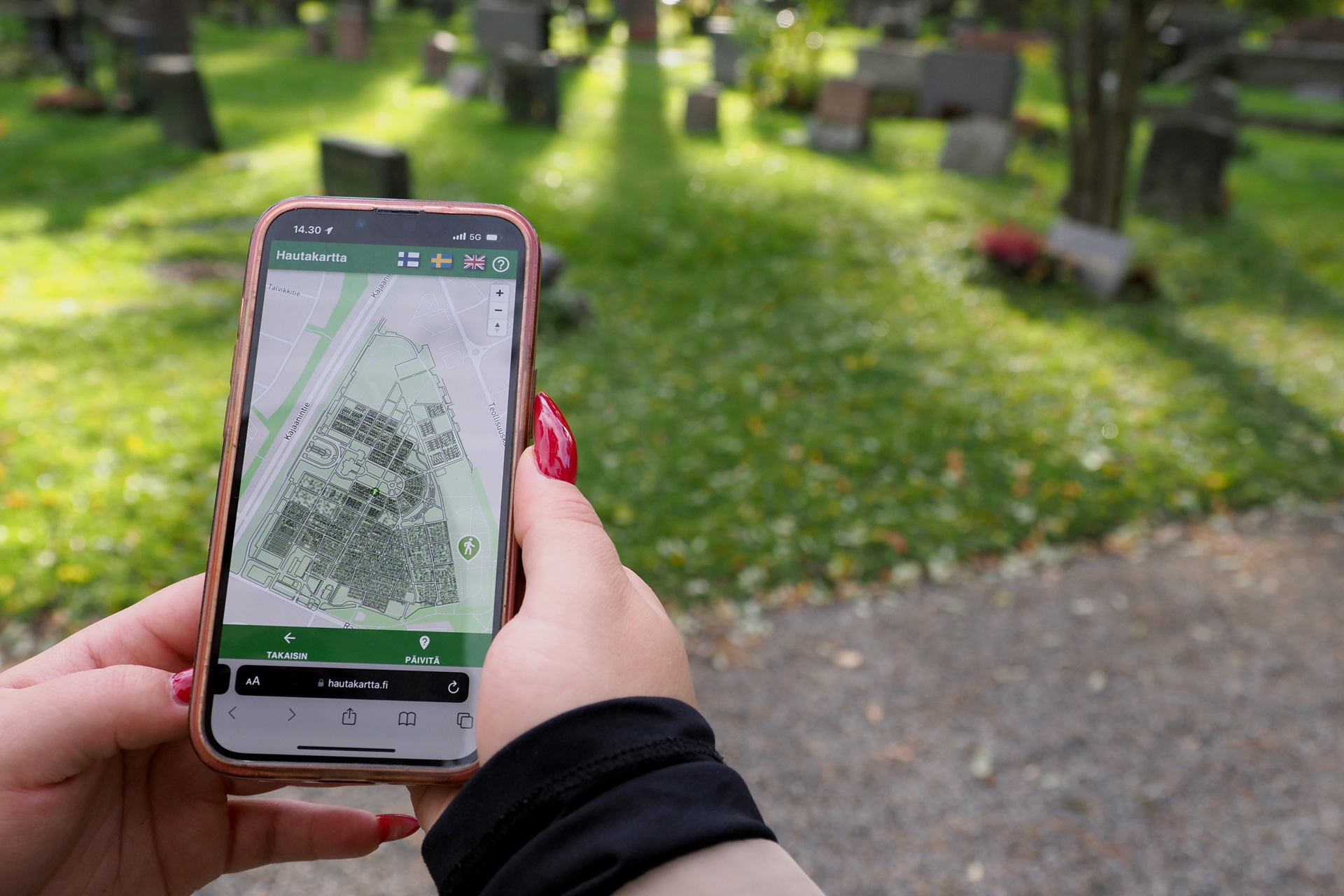Lähikuva käsissä olevasta puhelimesta, jonka näytöllä on kartta hautausmaasta, taustalla hautausmaata.