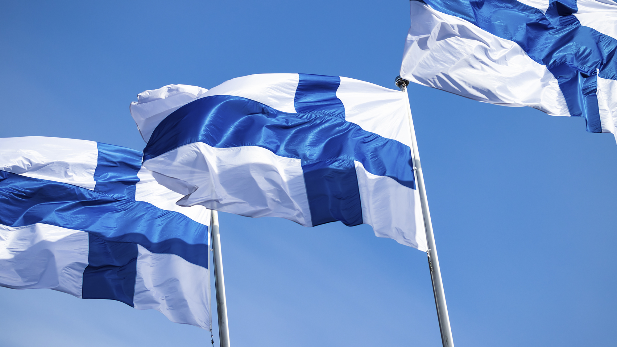Kolme Suomen lippua sinistä taivasta vasten.
